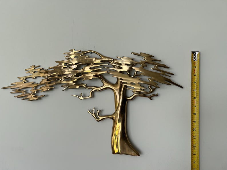 K&ouml;nnte beinhalten: Wandskulptur aus Messing in Form eines Baumes. Der Baum hat einen detaillierten Stamm und &Auml;ste mit einer strukturierten Krone. Die Skulptur ist etwa 35,5 cm hoch. Der Baum ist an einer hellgrauen Wand befestigt.