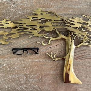 K&ouml;nnte beinhalten: Eine goldfarbene Metallbaumskulptur mit detailliertem Design und einer schwarzen Brille. Der Baum hat einen strukturierten Stamm und &Auml;ste, und die Bl&auml;tter sind aufwendig geschnitten. Die Brille hat einen schwarzen Rahmen.