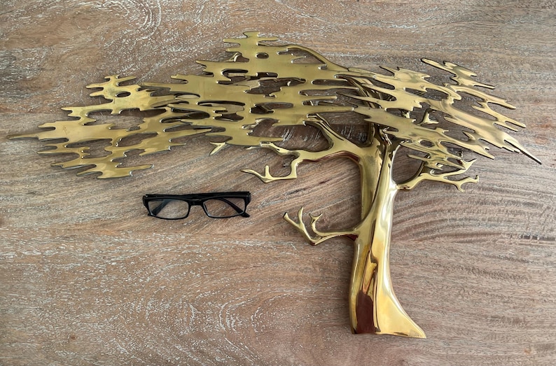 K&ouml;nnte beinhalten: Eine goldfarbene Metallbaumskulptur mit detailliertem Design und einer schwarzen Brille. Der Baum hat einen strukturierten Stamm und &Auml;ste, und die Bl&auml;tter sind aufwendig geschnitten. Die Brille hat einen schwarzen Rahmen.