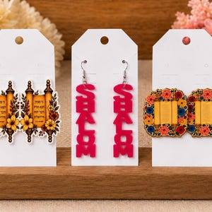 May include: Three pairs of earrings displayed on white cards. The left pair features yellow Torah scrolls with floral accents. The center pair consists of bright pink vertical dangles spelling SHALOM. The right pair shows square Torah scrolls with colorful floral borders.