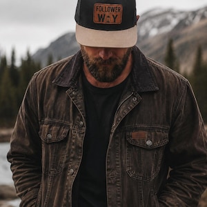 May include: A black baseball cap with a tan brim and a brown leather patch on the front featuring the text "FOLLOWER OF THE WAY" and a small lantern icon. The cap is worn with a dark brown distressed denim jacket over a plain black t-shirt.