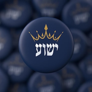 May include: A navy blue button with a gold crown design and white Hebrew text. The button is round and has a glossy finish. The background is blurred with similar buttons.