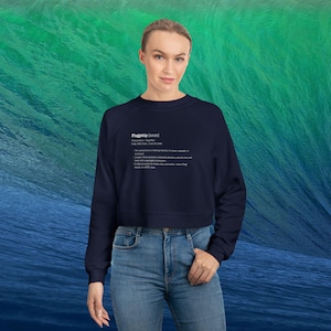 May include: A navy blue cropped sweatshirt with the word "Flagship" and its definition printed in white. The model is wearing blue jeans. The background is a gradient of green and blue.