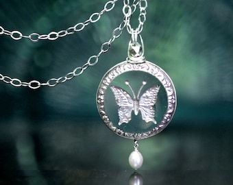 May include: A silver-toned circular pendant necklace featuring a detailed butterfly motif encased within a decorative frame. The pendant hangs from a delicate silver chain and includes a small, white, teardrop-shaped pearl accent at the bottom.