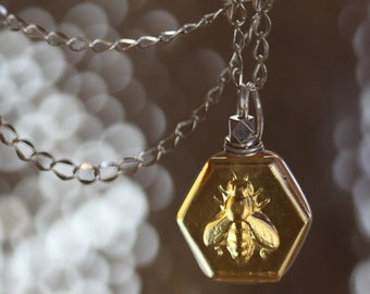 May include: A silver chain necklace with a hexagonal pendant featuring a bee encased in clear amber-colored resin.