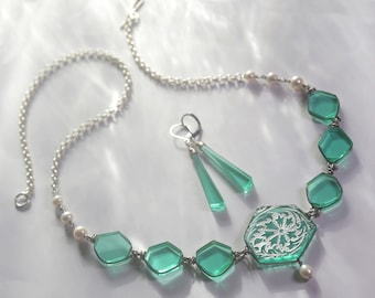 May include: A teal green glass necklace with a silver chain and white pearl accents. The necklace features a large hexagonal pendant with a white etched design. Matching teal green glass earrings are also included.