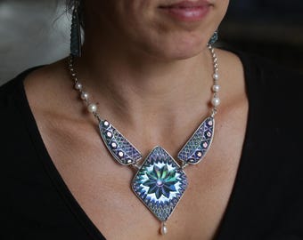 May include: A silver necklace with a large iridescent blue and purple pendant featuring a geometric design. The pendant is surrounded by smaller iridescent blue and purple pieces. The necklace is strung with white pearls.