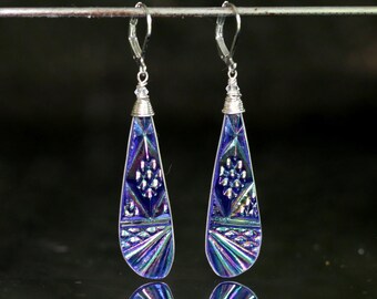 May include: A pair of drop earrings featuring elongated, teardrop-shaped components with a metallic, iridescent blue and purple finish. The surface displays geometric patterns, including a central floral motif, textured scales, and radiating lines.