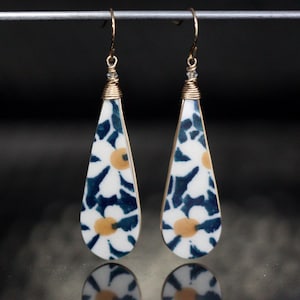May include: A pair of teardrop-shaped earrings with a white background and a blue and yellow floral pattern. The earrings are made with gold wire and have a small crystal at the top of the wire.