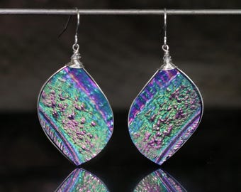May include: A pair of iridescent teardrop earrings. The earrings feature a textured, multi-colored surface with shades of green, purple, and blue. The edges are outlined in silver, and the hooks are silver. The earrings are suspended from a silver wire.
