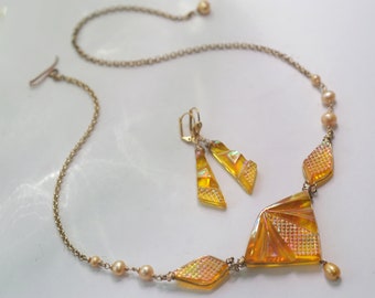 May include: A gold chain necklace with a large, iridescent yellow pendant and smaller matching earrings. The pendant is made of faceted glass and has a geometric design. The necklace is accented with small pearl beads.