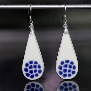 May include: A pair of teardrop-shaped earrings with a white ceramic base and a blue and white geometric pattern. The earrings are suspended from silver hooks.