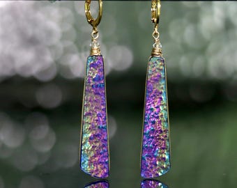 May include: A pair of long, trapezoidal drop earrings featuring iridescent, textured surfaces with vibrant purple, blue, and gold hues. Each earring is suspended from a gold-toned lever-back clasp by a small wire-wrapped loop at the top.