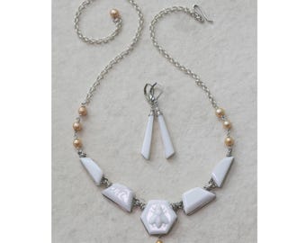 May include: A jewelry set consisting of a necklace and matching earrings. The necklace features a silver chain with peach-colored pearls, white geometric resin pieces, and a central hexagonal pendant with a bee motif. The earrings are white.