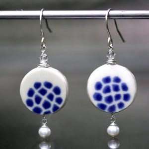 May include: A pair of white ceramic earrings with a blue and white geometric pattern. Each earring has a small pearl drop.