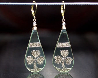 May include: A pair of gold dangle earrings with a clear teardrop-shaped glass charm featuring a silver shamrock design.