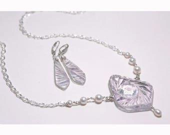 May include: A silver necklace and matching earrings set. The necklace features a square, carved pendant with a pearl drop, and is accented with small pearls. The earrings are long, carved, and dangle. The jewelry is set against a white background.