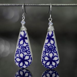 May include: A pair of teardrop-shaped earrings featuring a white ceramic base with a blue floral pattern. Each earring is topped with a small clear crystal bead and silver-toned wire wrapping, suspended from silver-toned metal ear hooks.