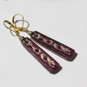 Op de afbeelding: Een paar oorhangers met gouden haakjes en paarse glazen druppels. De glazen druppels hebben een bloemmotief dat in goud is gegraveerd.