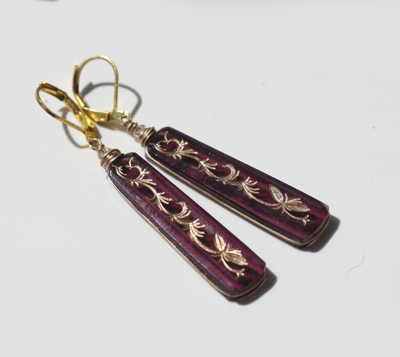 Op de afbeelding: Een paar oorhangers met gouden haakjes en paarse glazen druppels. De glazen druppels hebben een bloemmotief dat in goud is gegraveerd.