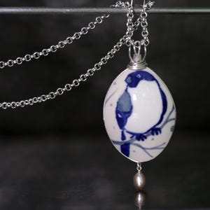 May include: A silver necklace with a white and blue porcelain pendant. The pendant is oval-shaped and features a blue bird perched on a branch. A small, gray pearl hangs from the bottom of the pendant. The necklace chain is also silver.