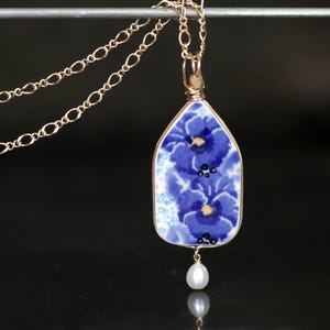 May include: A gold-toned necklace featuring a pendant with a blue floral design. The pendant has a teardrop shape and is adorned with a small pearl at the bottom. The chain is delicate and has a link design.
