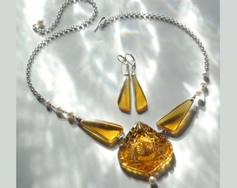 May include: A silver chain necklace with a large amber pendant and two smaller amber drop earrings. The pendant is a geometric shape with a textured surface. The necklace is accented with small white pearls.