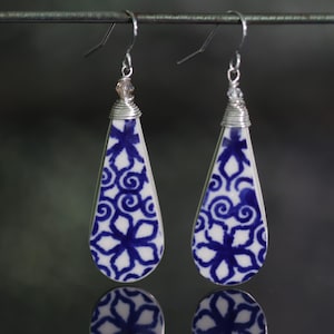 May include: A pair of teardrop-shaped earrings featuring a white and blue floral pattern. The ceramic pieces are wrapped in silver-toned wire at the top, accented with a small clear bead, and hang from silver-toned metal ear wires.