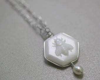 May include: A white hexagonal pendant necklace with a carved bee design and a single white pearl drop. The necklace is made of silver chain.