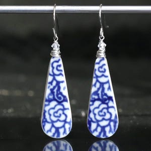 May include: A pair of teardrop-shaped earrings with a blue and white floral pattern. The earrings are made from porcelain and have silver wire wrapped around the top with a crystal bead.