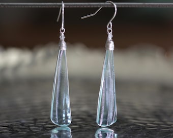 May include: A pair of elegant, teardrop-shaped earrings. Each earring features a clear, light blue glass pendant suspended from a silver-colored hook. The glass has a textured surface and is wrapped with silver wire at the top.