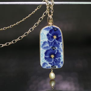 May include: Gold-tone necklace with a rectangular pendant featuring blue floral designs on a white background. A small, teardrop-shaped pearl dangles from the bottom of the pendant. The chain is delicate and has a double strand design.