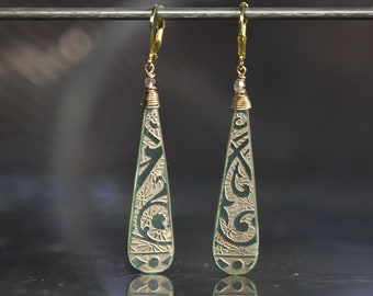May include: A pair of gold dangle earrings with green teardrop-shaped pendants. The pendants have a detailed etched design.