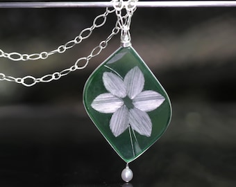 May include: A green glass pendant with a white flower design and a pearl hanging from the bottom.