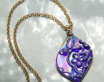 May include: A gold-colored chain necklace with a large, iridescent pendant. The pendant is a deep purple color with floral designs and a gold-colored border. The necklace is displayed on a light-colored surface.