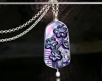 May include: A silver necklace featuring a pendant with a floral design. The pendant has a pearlescent, iridescent finish with shades of purple, blue, and pink. A small, white pearl dangles from the bottom of the pendant.