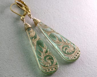 May include: A pair of elegant, teardrop-shaped earrings. The earrings feature a light green, translucent design with gold-colored filigree patterns. The gold-colored ear wires and wire wrapping add to the jewelry's delicate appearance.
