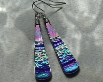 May include: A pair of iridescent dangle earrings. The earrings feature a long, slender, rectangular shape with a rounded bottom. The colors shift from pink to blue and green. The ear wires are silver-toned.