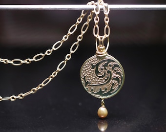 May include: Gold-toned necklace featuring a circular pendant with an intricate, embossed design and a small, pear-shaped pearl drop. The chain has a delicate, linked pattern. The pendant has a dark green background.