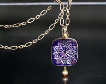 May include: A gold chain necklace with a square pendant made of iridescent purple glass with a cut-glass design. A small pearl dangles from the bottom of the pendant.