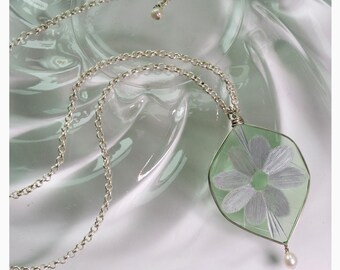 May include: A silver chain necklace with a green glass pendant featuring a white embroidered flower. The pendant is attached to a silver chain with a pearl at the bottom.