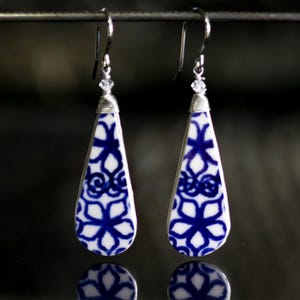 May include: A pair of teardrop-shaped earrings with a blue and white porcelain design. The earrings are made with silver wire and have a small crystal accent.