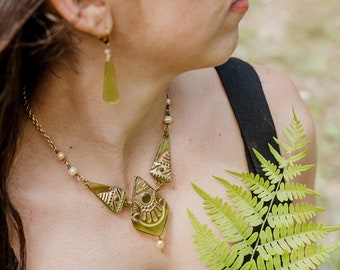 May include: A gold necklace with a green glass pendant featuring intricate designs. The pendant is made up of three pieces, each with a different pattern. The necklace also features small pearl beads. The necklace is worn by a person with long brown hair and a black tank top.