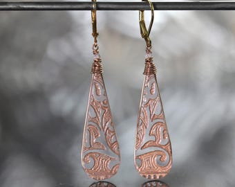May include: A pair of copper-colored teardrop earrings with a floral design. The earrings have a light gray background and are suspended from gold-colored ear wires. The design is etched into the metal, creating a detailed pattern.
