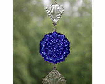 May include: A blue glass suncatcher with a clear glass diamond shape above and below it. The suncatcher is hanging from a silver chain.