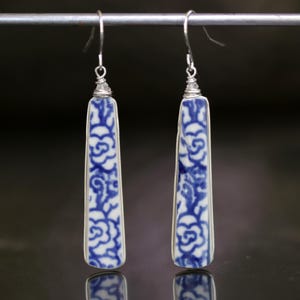 May include: A pair of long, rectangular earrings with a blue and white floral pattern. The earrings have a silver-colored metal frame and hook. The design is reminiscent of porcelain. The earrings are hanging from a silver bar.