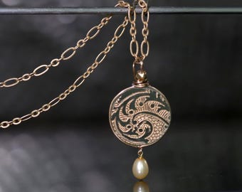 May include: Gold-toned pendant necklace featuring a circular pendant with an etched design and a dangling pearl. The necklace has two delicate chains. The pendant's design is intricate, with swirling patterns and a small pearl drop.