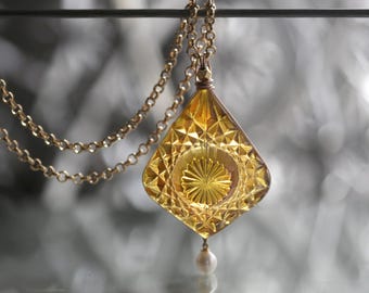 May include: A gold chain necklace with a large, faceted, amber-colored glass pendant. The pendant has a diamond-shaped design with a smaller, round pearl hanging from the bottom.