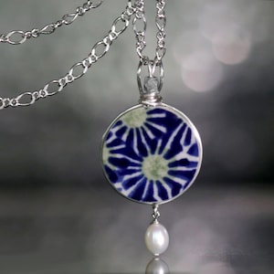 May include: A silver chain necklace with a round pendant featuring a blue and white floral design. A single white pearl dangles from the bottom of the pendant.