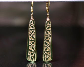 May include: A pair of gold dangle earrings with green glass teardrop-shaped pendants. The pendants have a gold geometric pattern.